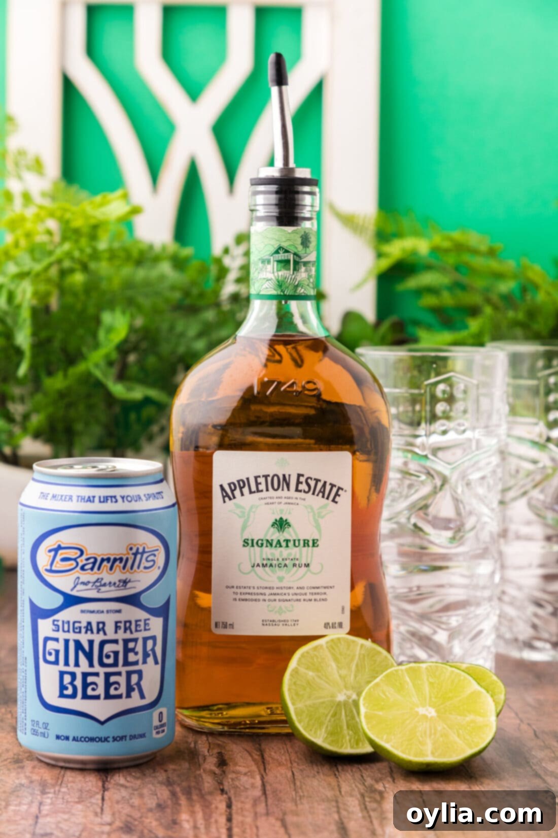 A flat lay photograph of the fresh ingredients for a Jamaican Mule: a bottle of rum, ginger beer, limes, and a measuring jigger.