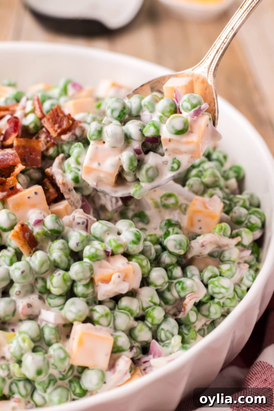 A serving spoon lifting a generous portion of creamy Pea Salad from a large white bowl, showcasing its rich texture and a mix of peas, bacon, and cheese