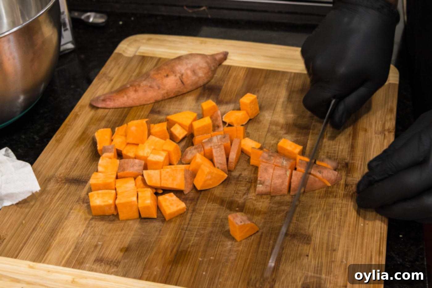 Dicing fresh sweet potatoes into uniform bite-sized chunks on a cutting board using a chef's knife.