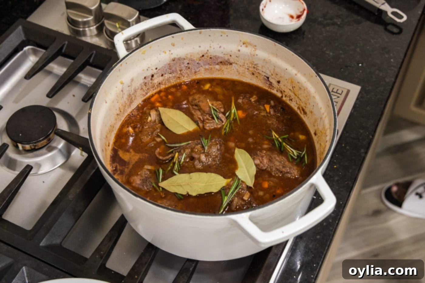 Braised beef short ribs simmering in a Dutch oven, with fresh rosemary and bay leaves visible in the rich red wine sauce.