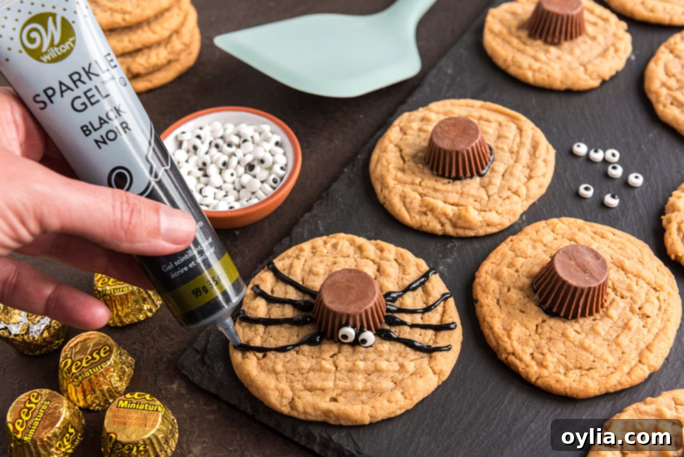 Piping black decorator gel onto a spider cookie to form the legs, starting from the Reese's cup body.