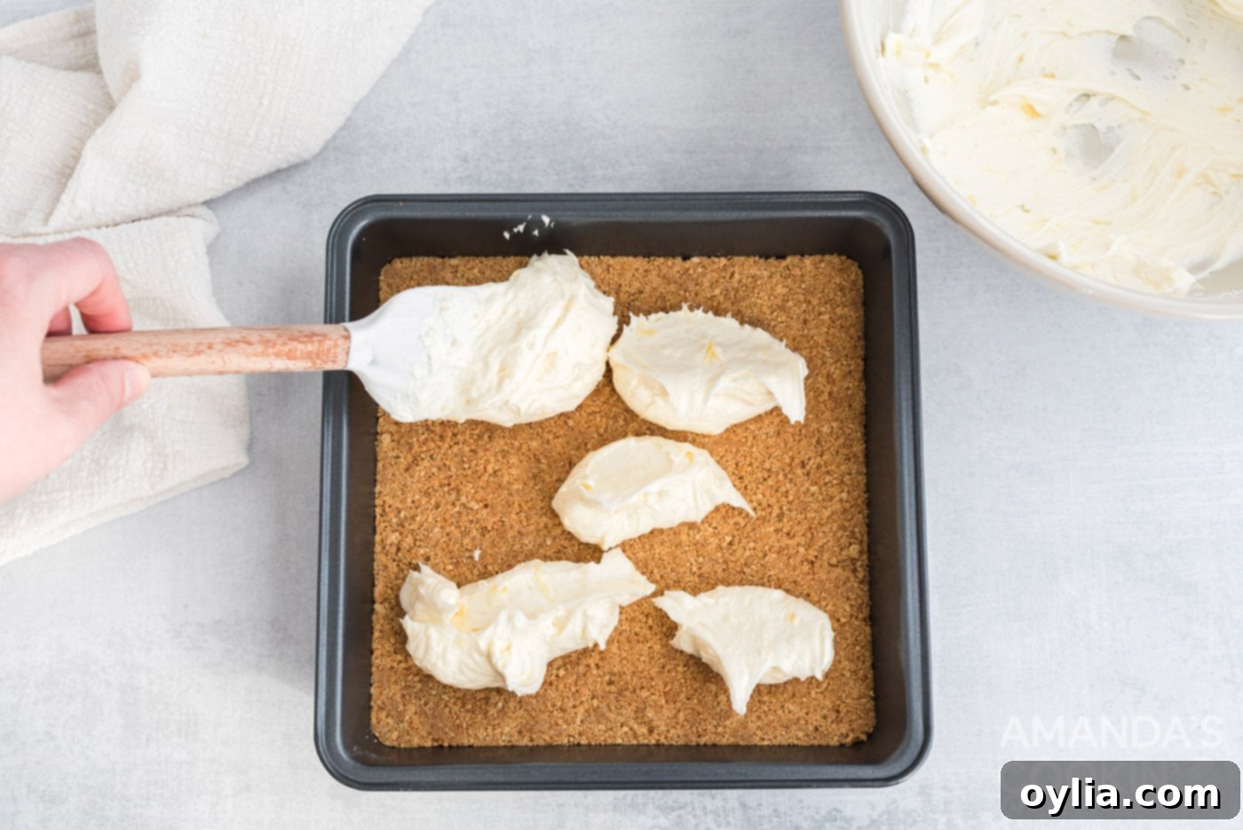 The smooth cream cheese and pineapple mixture being spread evenly over the graham cracker crust in the baking pan.