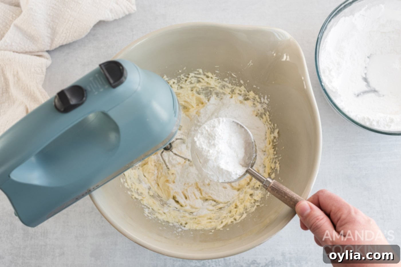 Powdered sugar being added to the creamy mixture of cream cheese and butter in a mixer bowl.