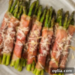 Close up photo of Prosciutto Wrapped Asparagus on a plate topped with shaved parmesan, perfectly golden and tender