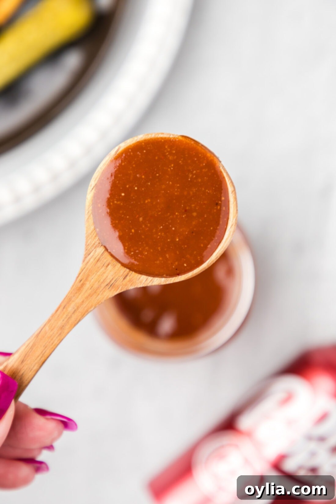 A spoon with Dr Pepper Barbecue Sauce held above a jar, showing its rich, glossy consistency.