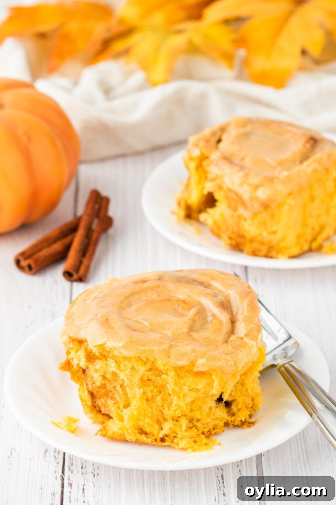 Two Homemade Pumpkin Cinnamon Rolls on plates, generously topped with pumpkin cream cheese frosting.