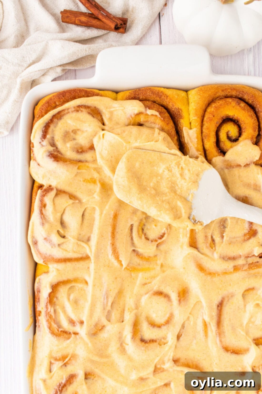Smooth pumpkin cream cheese frosting being generously spread onto baked pumpkin cinnamon rolls in a pan.