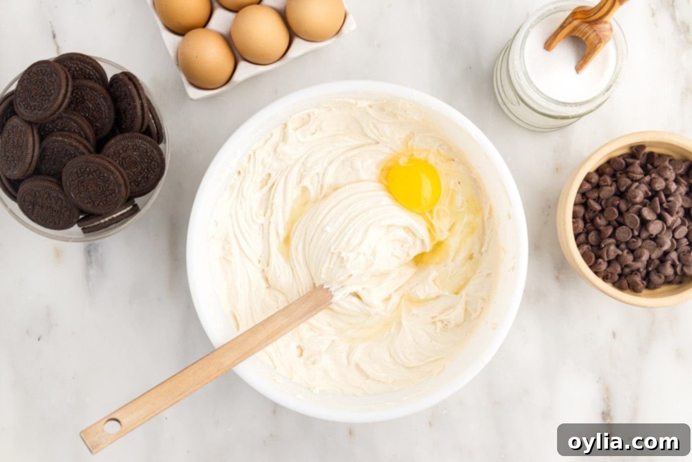 A large egg being gently added to the creamy cheesecake batter in a mixing bowl, to be incorporated by hand.