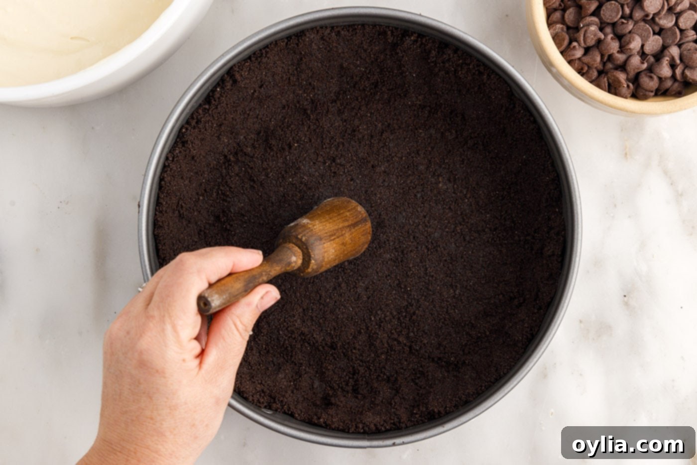 The Oreo cookie crumb mixture being pressed firmly into the bottom and up the sides of a springform pan to form a thick crust.