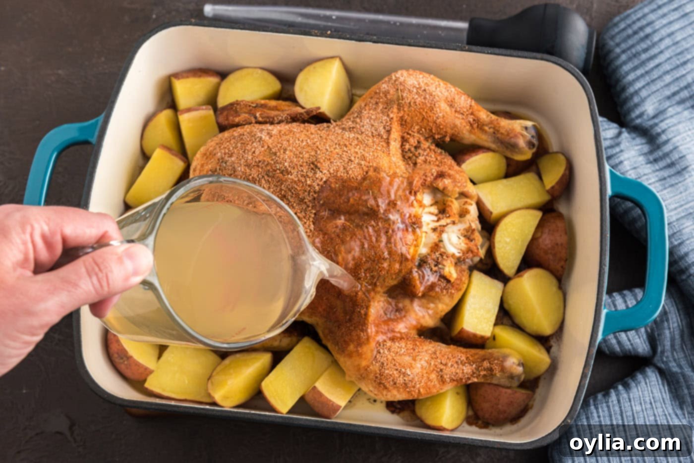 Pouring chicken broth over a whole roasted chicken in a pan with potatoes