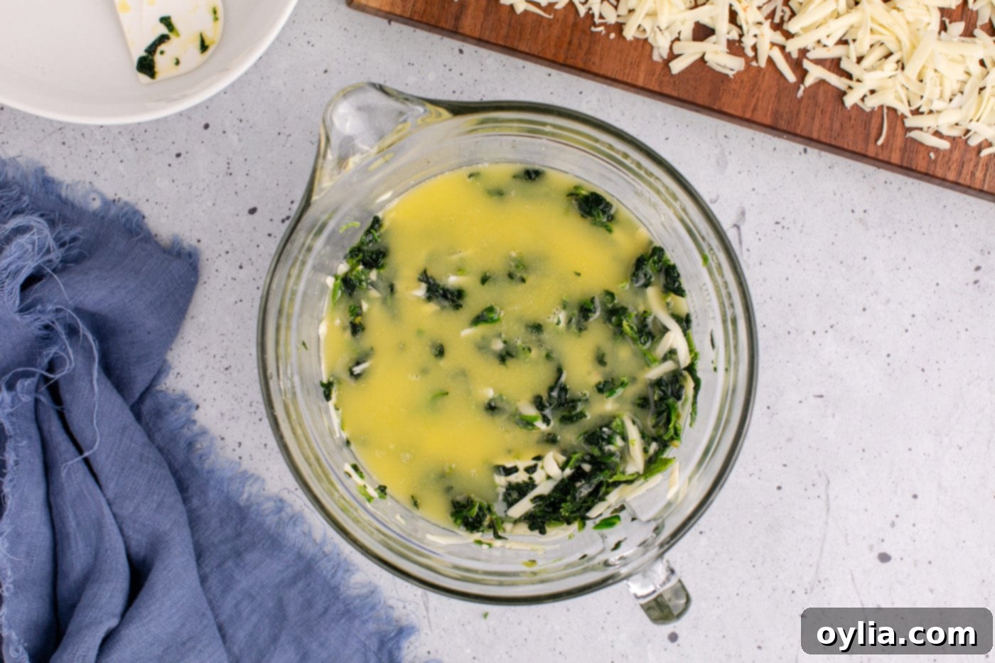 melted butter being poured over the mixed spinach souffle ingredients in a bowl, creating a glossy layer before final mixing.