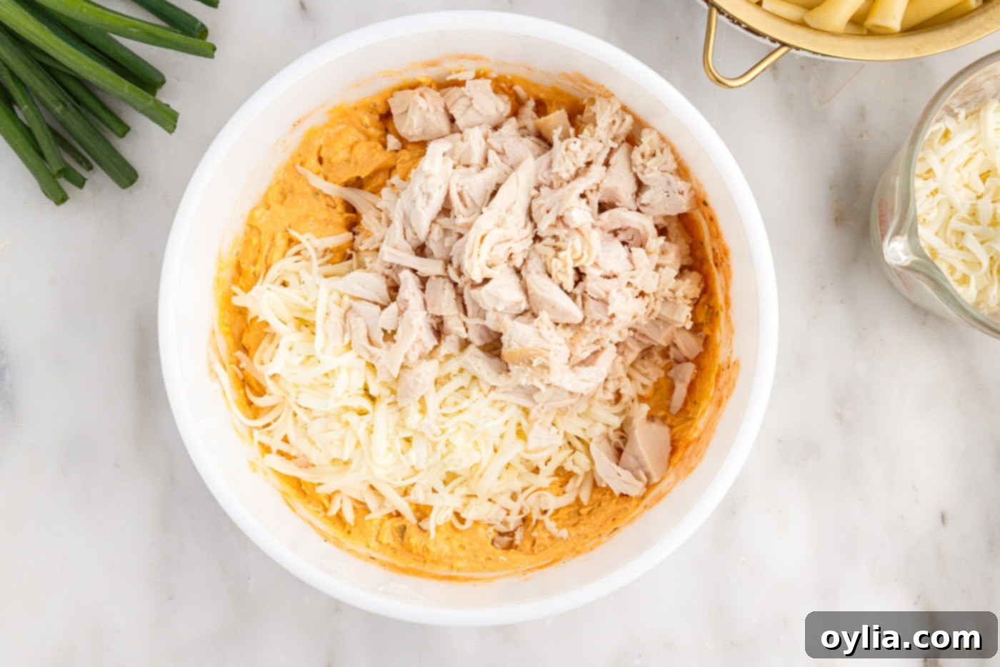 Adding shredded mozzarella cheese and cooked chicken to the buffalo chicken sauce mixture in a bowl.