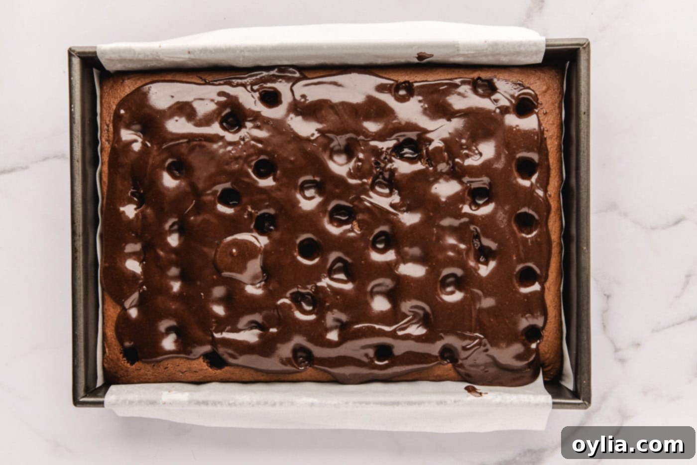Warm fudge sauce being poured and spread over the poked chocolate cake, sinking into the holes.