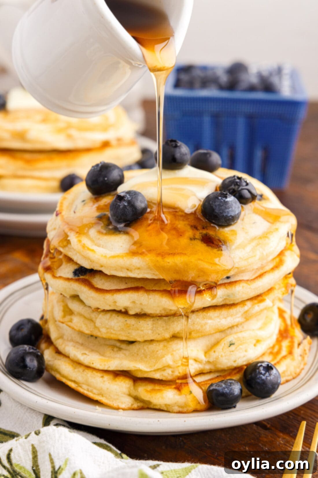 Berry Burst Pancakes 2 Syrup being poured generously over a towering stack of thick, fluffy blueberry pancakes, promising a sweet and satisfying breakfast experience.