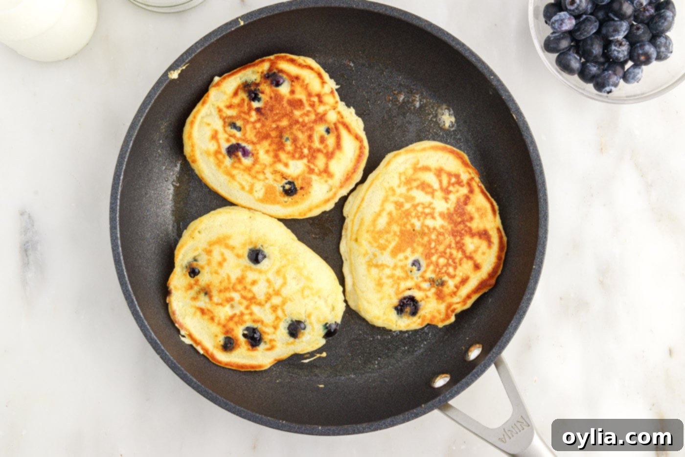 Cooked blueberry pancakes in a skillet, showing one side golden brown and ready to be flipped, while the other cooks.