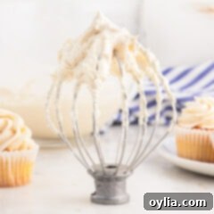 Brown Butter Frosting on a whisk attachment for mixer with cupcakes behind it, ready to be spread or piped.