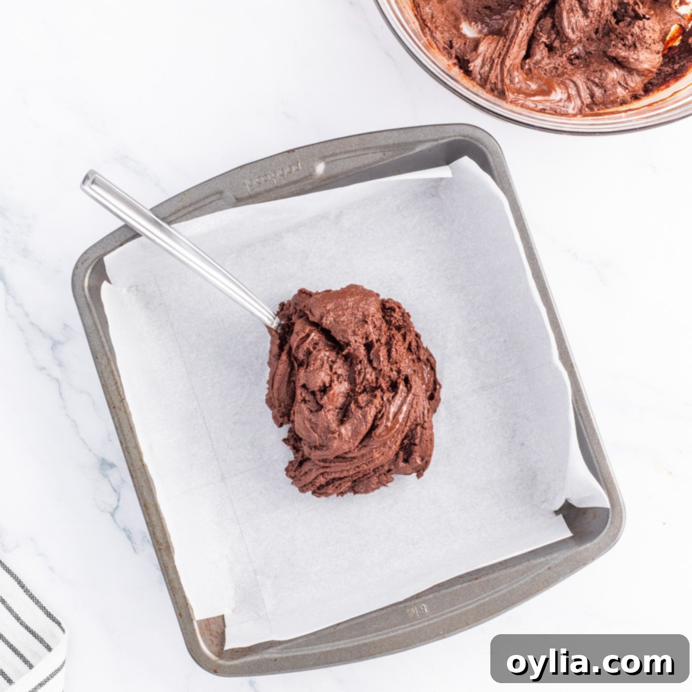 Spreading smooth chocolate fudge mixture evenly into a parchment-lined baking pan, forming the first layer