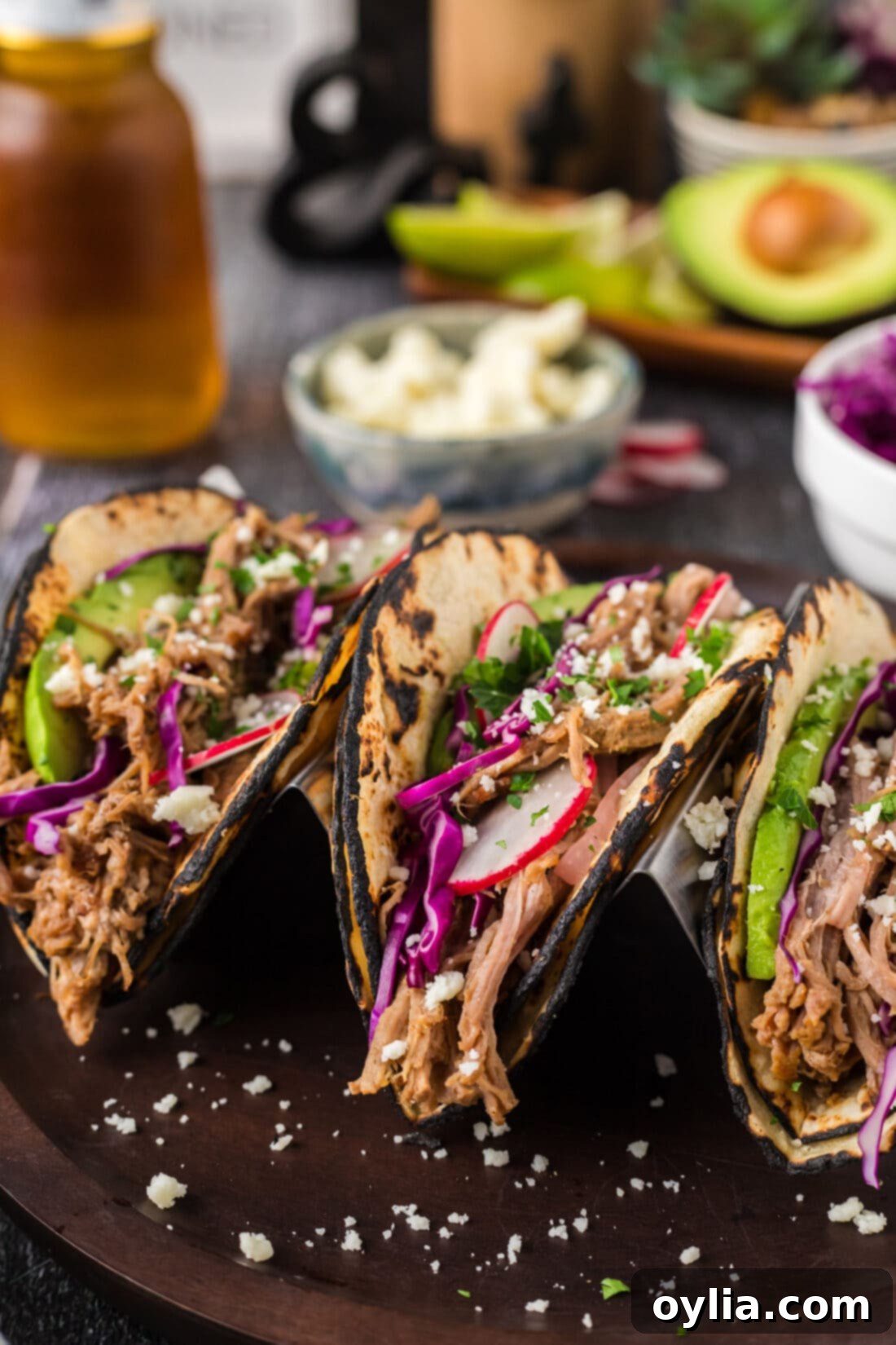 Ultimate Pulled Pork Tacos 2 Close up photo of three Pulled Pork Tacos on a plate, garnished with fresh red cabbage, cotija cheese, and a lime wedge