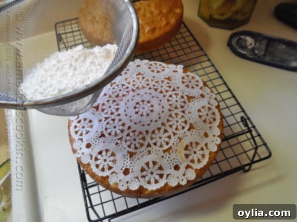 Powdered sugar being elegantly sprinkled through a paper doily placed on top of a Merryfield apple cake, creating a decorative pattern.