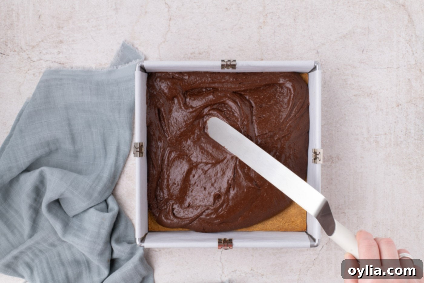 Spreading thick, glossy brownie batter evenly over the baked graham cracker crust in a baking pan.