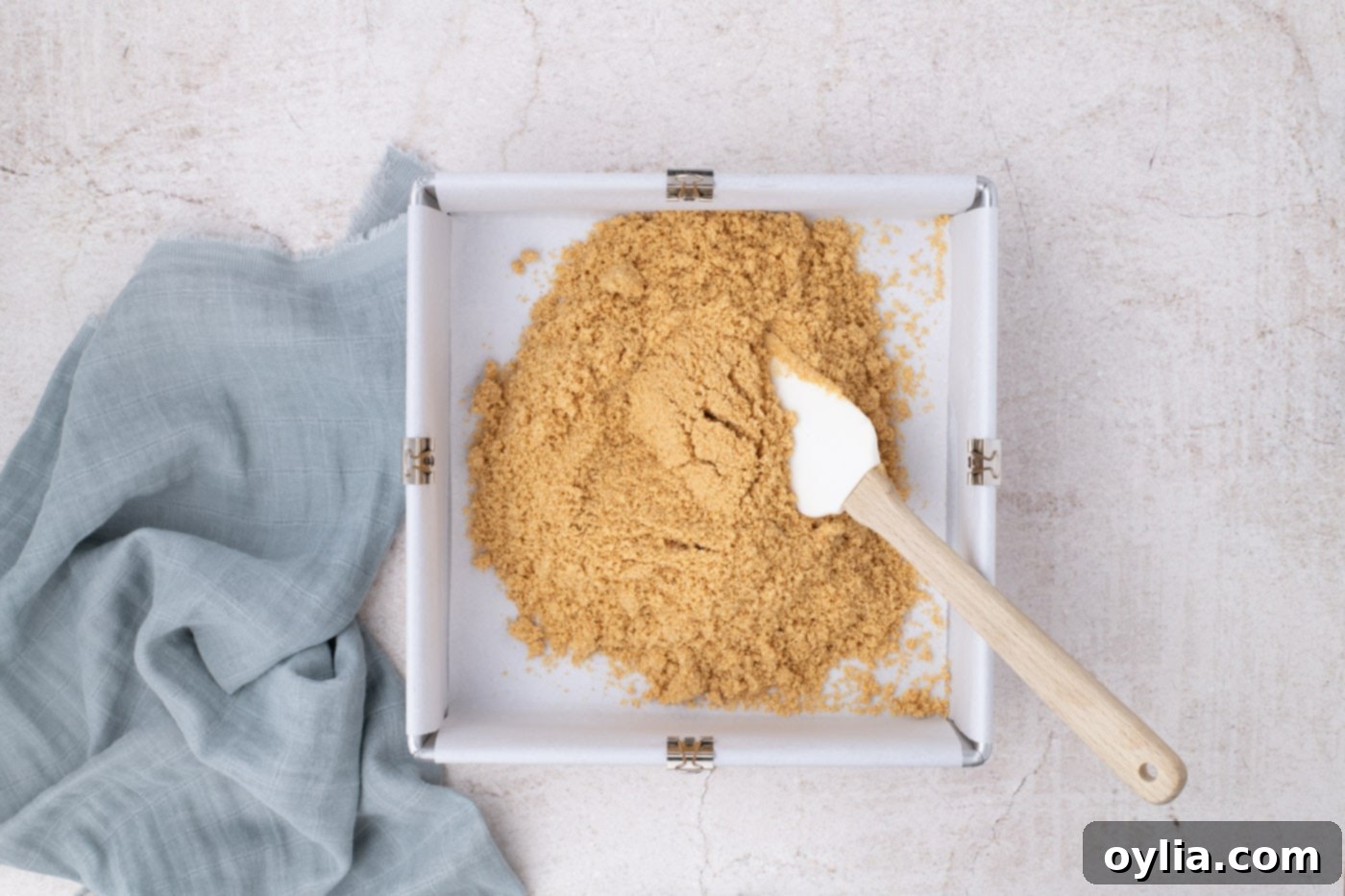 Spreading graham cracker crust evenly on the bottom of a parchment-lined 9x9 inch baking pan, ready for baking.