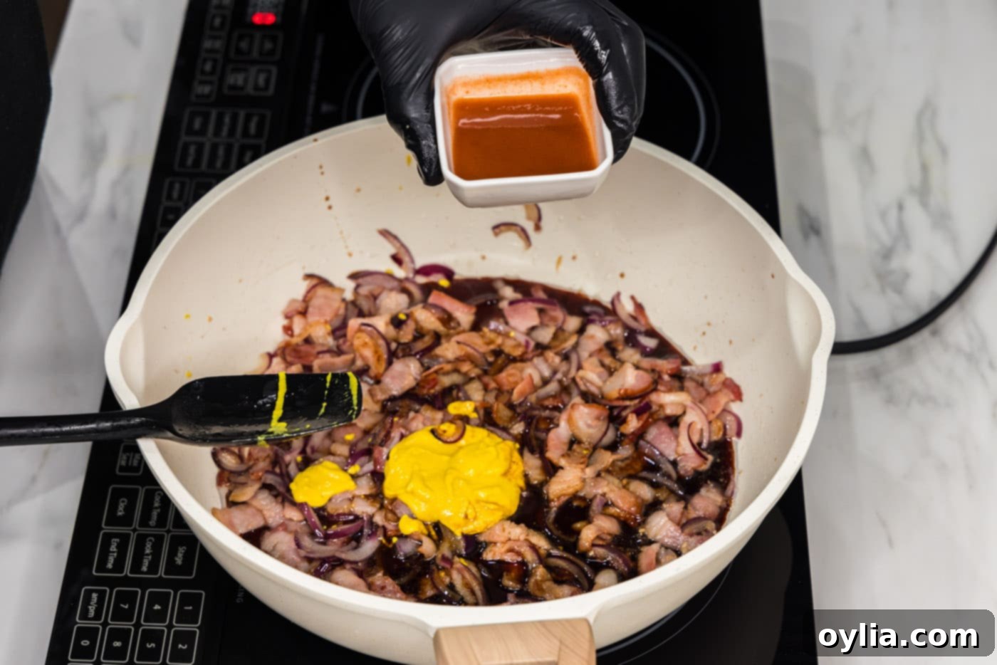 adding hot sauce to mixture of bacon, mustard, onions, and soy sauce in a skillet for a spicy kick.