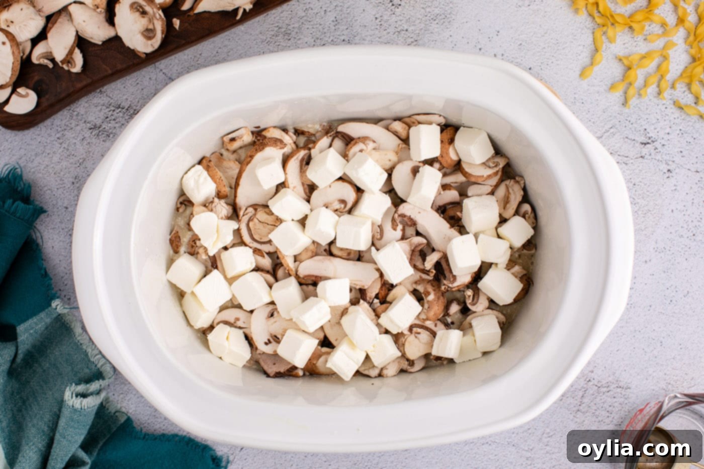 Cubed softened cream cheese placed on top of the chicken and mushroom mixture in the slow cooker, ready for cooking.