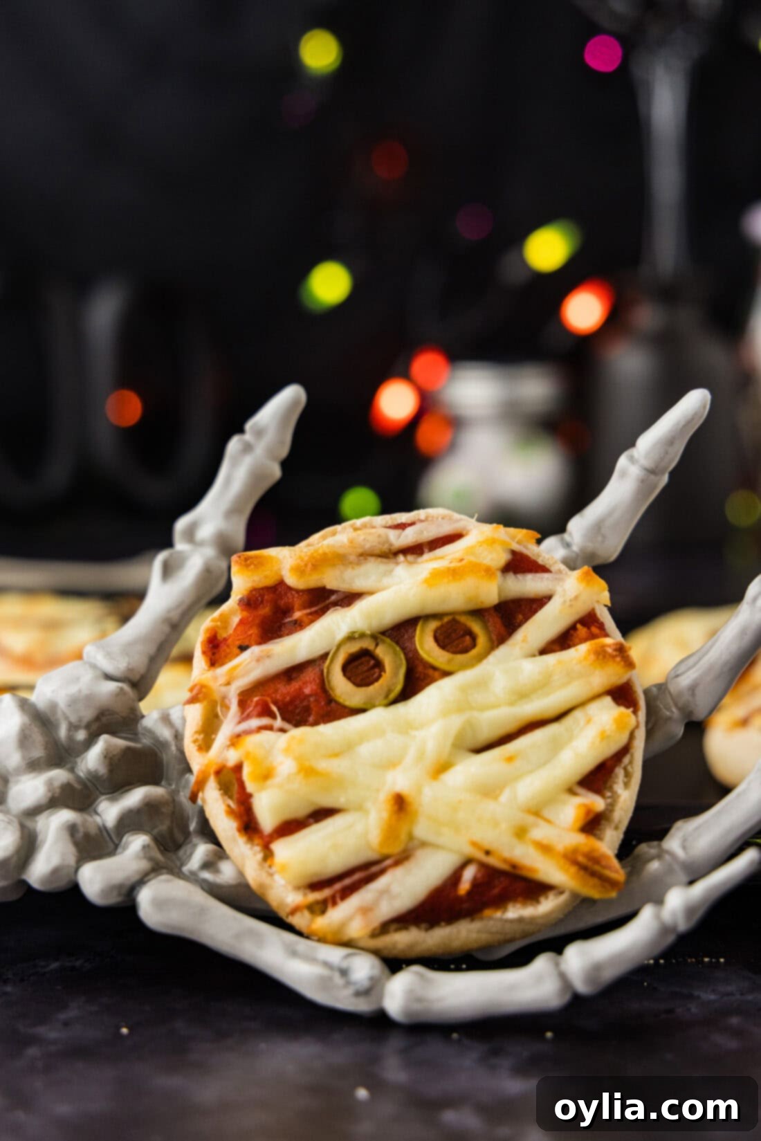 Mummy Pizza Bites 2 Mini Mummy Pizza held by a skeleton hand, showcasing the cheesy bandages and olive eyes, perfect for a Halloween party appetizer.