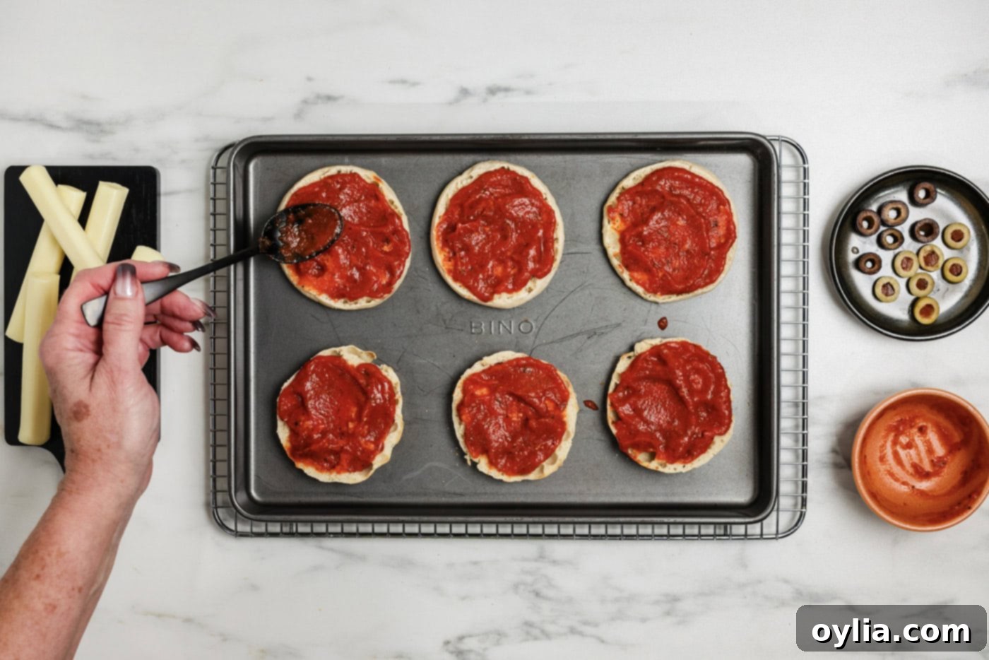 Mummy Pizza Bites 6 A close-up of English muffins being basted with bright red pizza sauce, preparing them for the mummy transformation.
