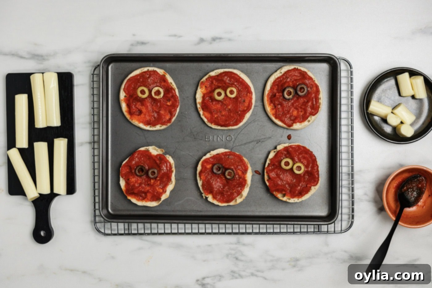 Mummy Pizza Bites 7 Sauced English muffins with sliced black olives strategically placed at the top to resemble spooky eyes, ready for the cheese bandages.