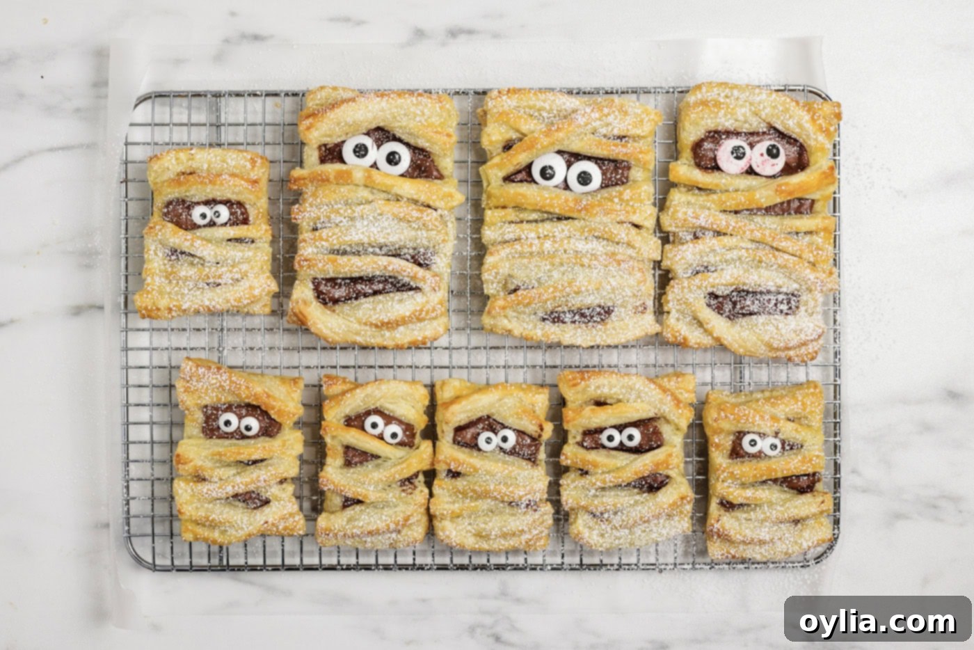 Bandaged Pastry Pockets 12 Powdered sugar being lightly dusted over freshly baked mummy hand pies arranged on a wire cooling rack, creating a ghostly effect.