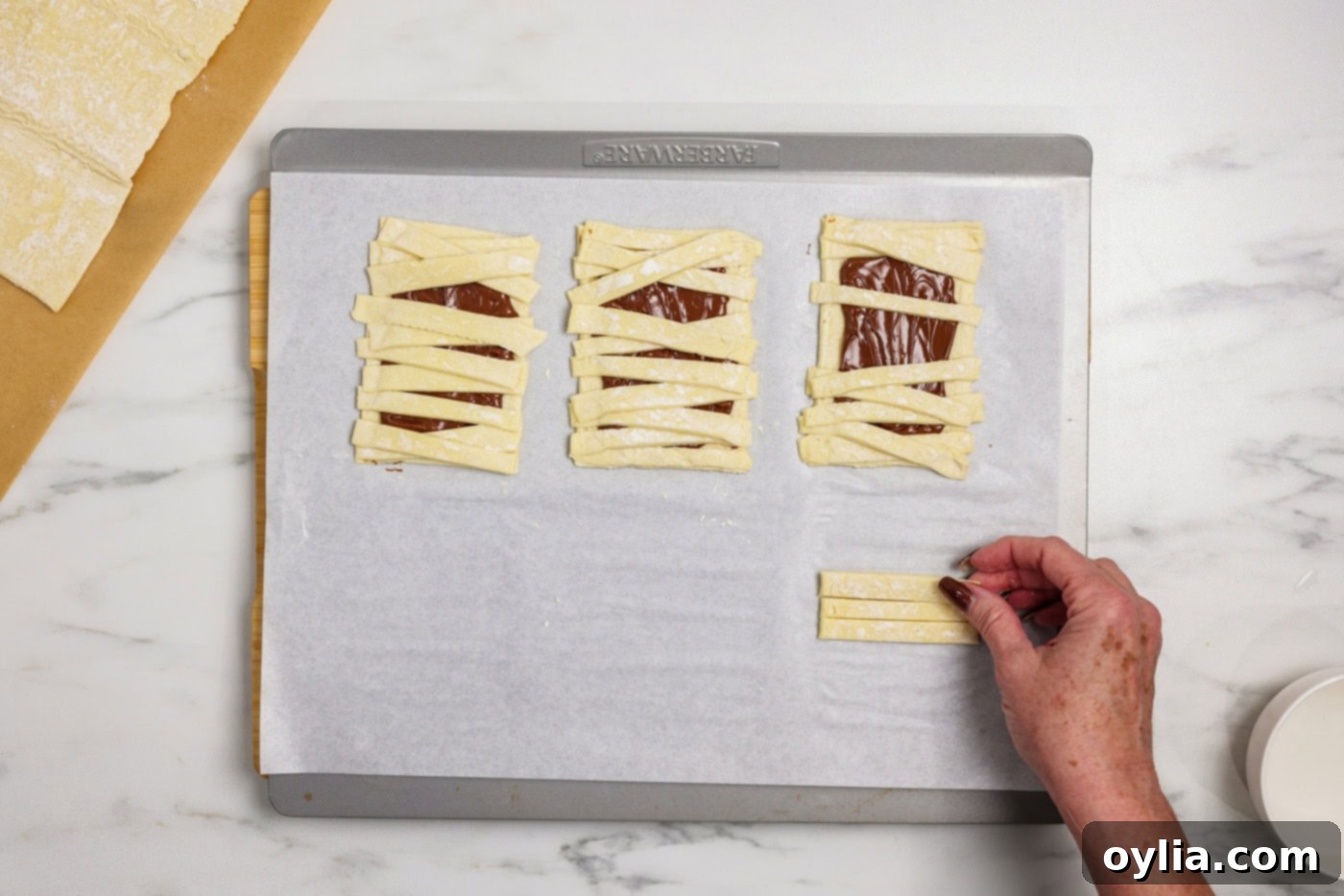 Bandaged Pastry Pockets 8 A hand gently arranging strips of puff pastry over a Nutella-coated rectangle, creating the layered look of mummy bandages.