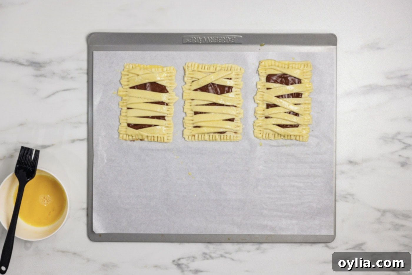 Bandaged Pastry Pockets 10 Mummy hand pies neatly arranged on a baking sheet, brushed with a prepared egg wash for a perfect golden finish.