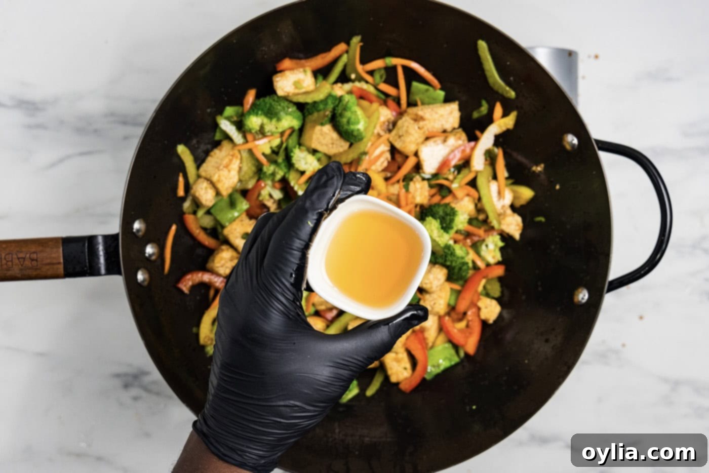 Flavorful Tofu Stir-Fry 8 hand holding a small bowl of sesame oil over tofu stir fry in a wok