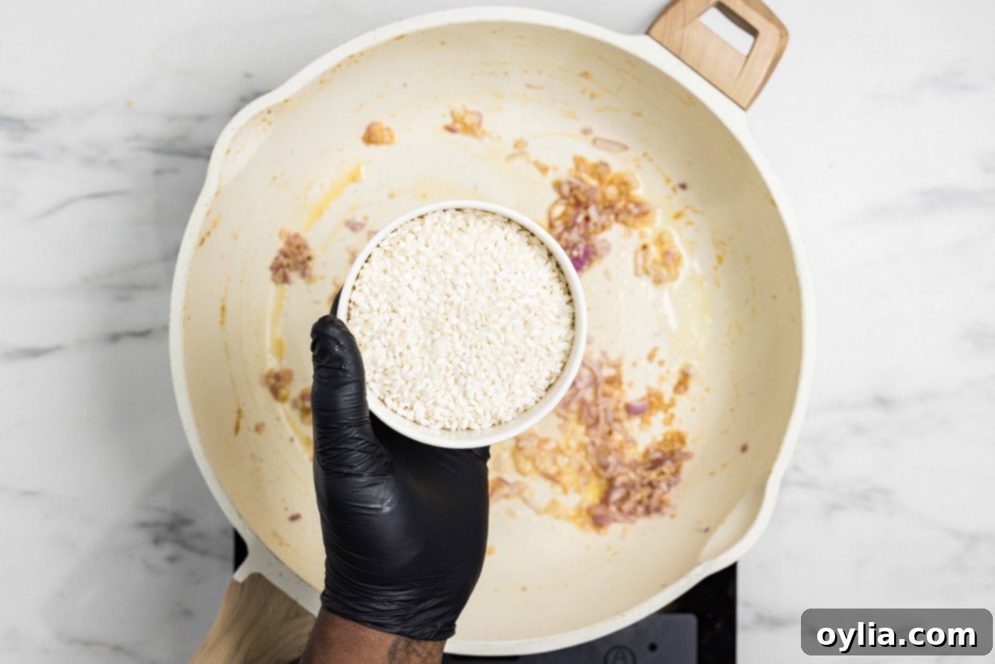 A hand holding a bowl of Arborio rice poised over a skillet containing sautéed garlic and shallots, ready to be added for toasting.