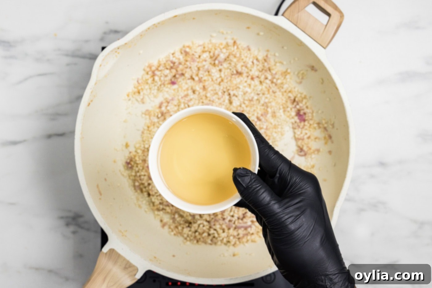 A hand holding a bowl of dry white wine as it is poured into a skillet with risotto rice, garlic, and shallots, to deglaze the pan.
