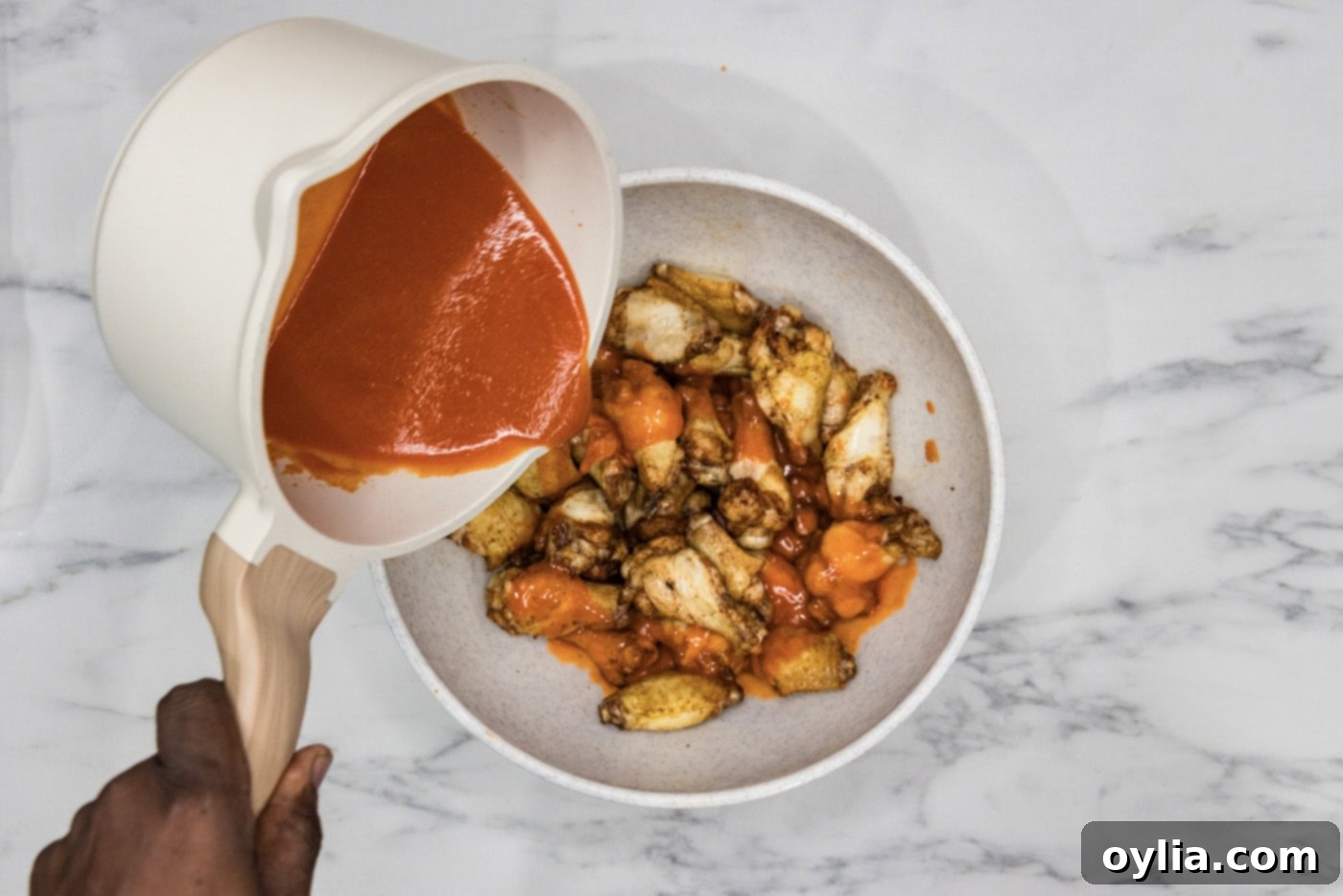 Homemade buffalo sauce being generously poured over a bowl of freshly fried chicken wings.