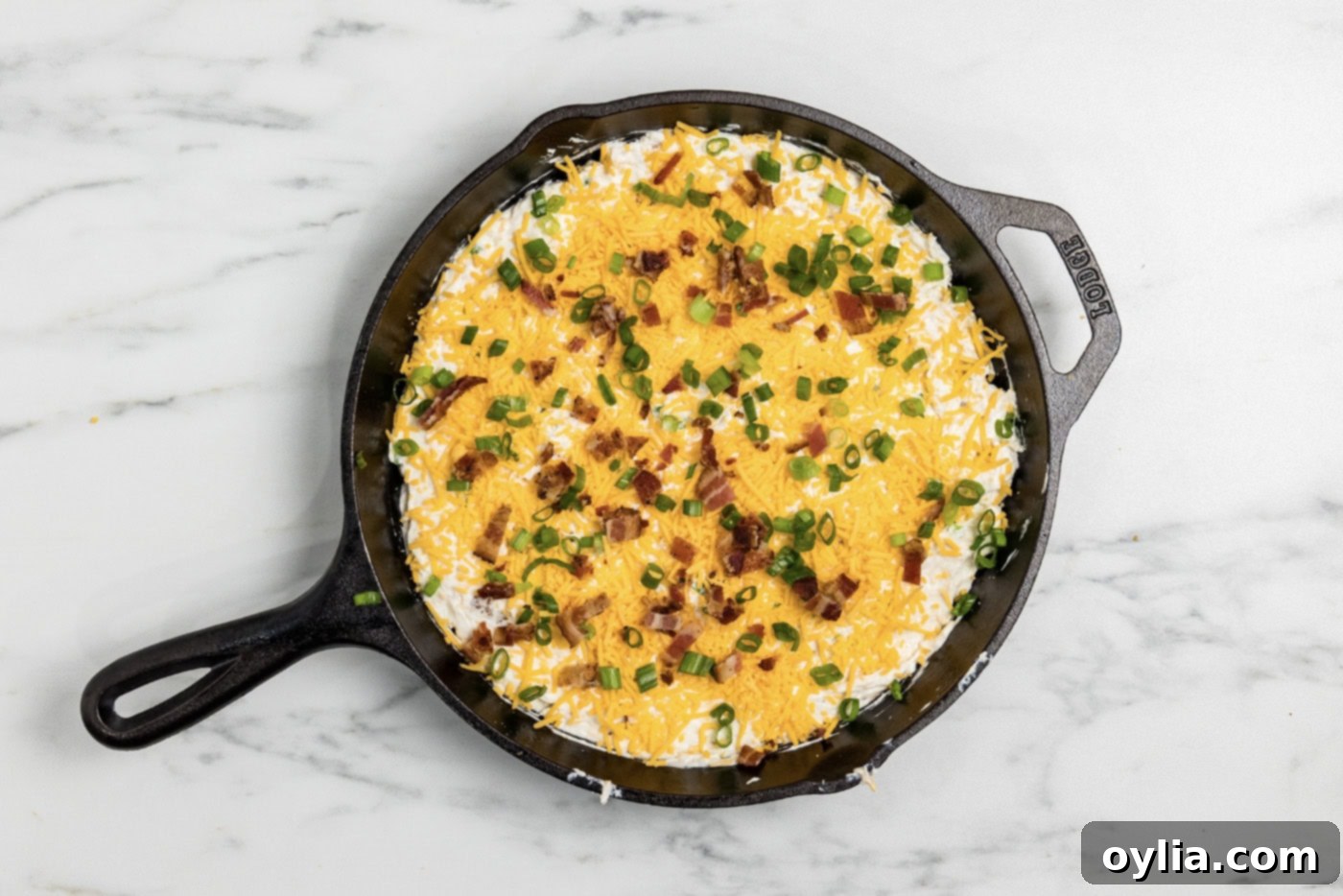 Dip covered with shredded cheddar, crumbled bacon, and sliced green onions in a cast iron skillet - Baking to cheesy perfection!